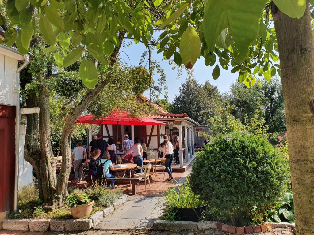 Eine Gruppe von Menschen versammelt sich an einem sonnigen Tag vor einem Café mit einem roten Sonnenschirm. Mehrere Personen sitzen an Holztischen im Freien und genießen die angenehme Atmosphäre.