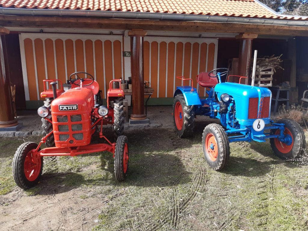 Zwei alte Traktoren, einer rot und einer blau, stehen nebeneinander vor einem Gebäude. Der rote Traktor trägt die Aufschrift „FAHR“. Der blaue Traktor hat die Nummer „6“ auf der Seite.